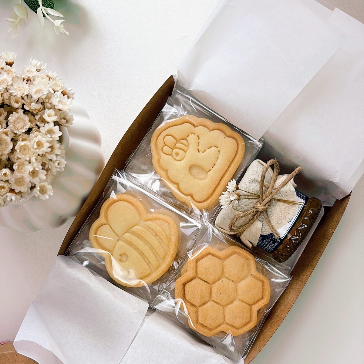 Caixa de presente com biscoitos amanteigados carimbados nos formatos de abelha, colmeia e favo de mel, embalados individualmente ao lado de um pote de mel