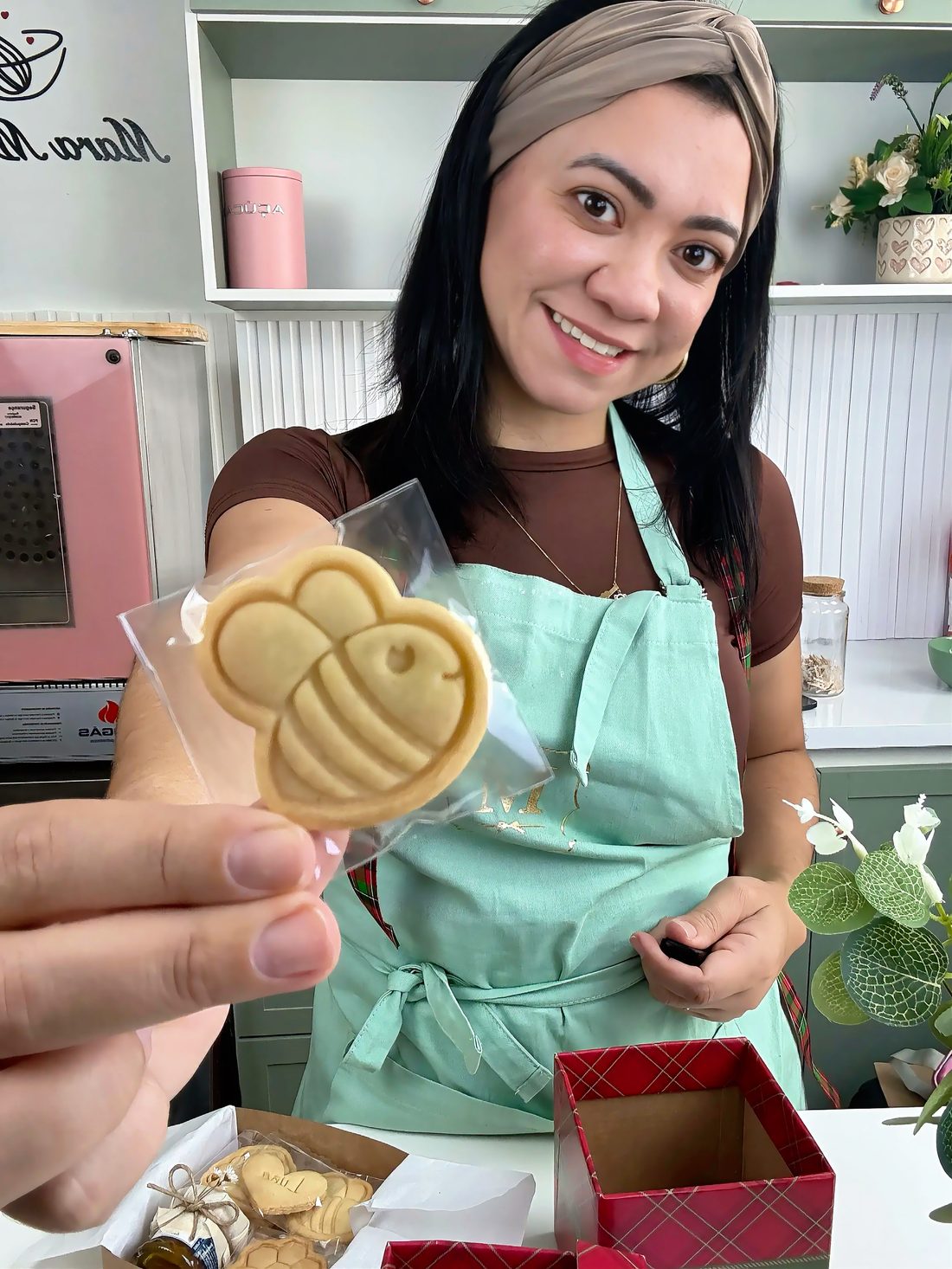Mara Morais, de avental verde-menta, segurando um biscoito amanteigado carimbado no formato de abelha em sua cozinha